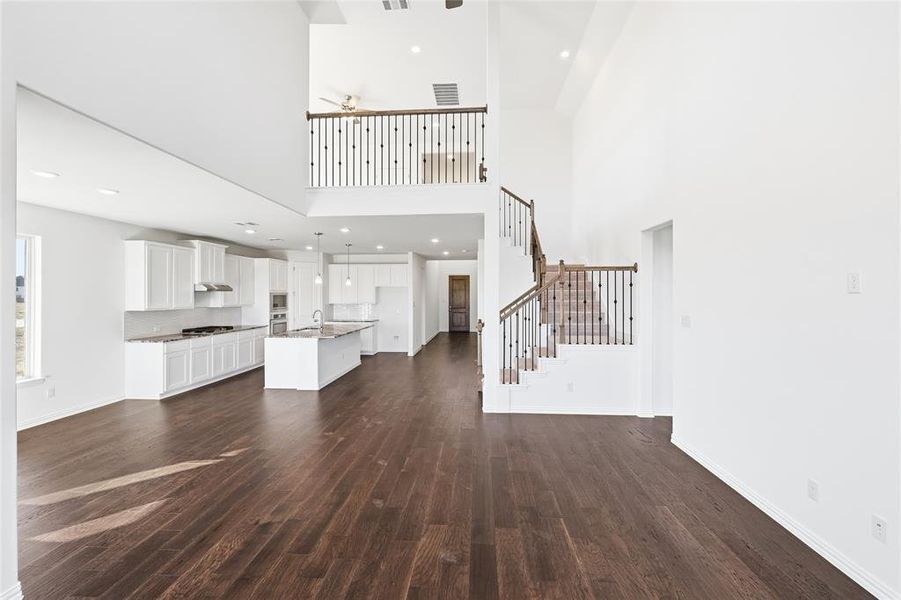 Expansive open-concept interior featuring high ceilings, wood-finish flooring, a contemporary kitchen with white cabinetry and an island, and a prominent staircase with wrought iron balusters