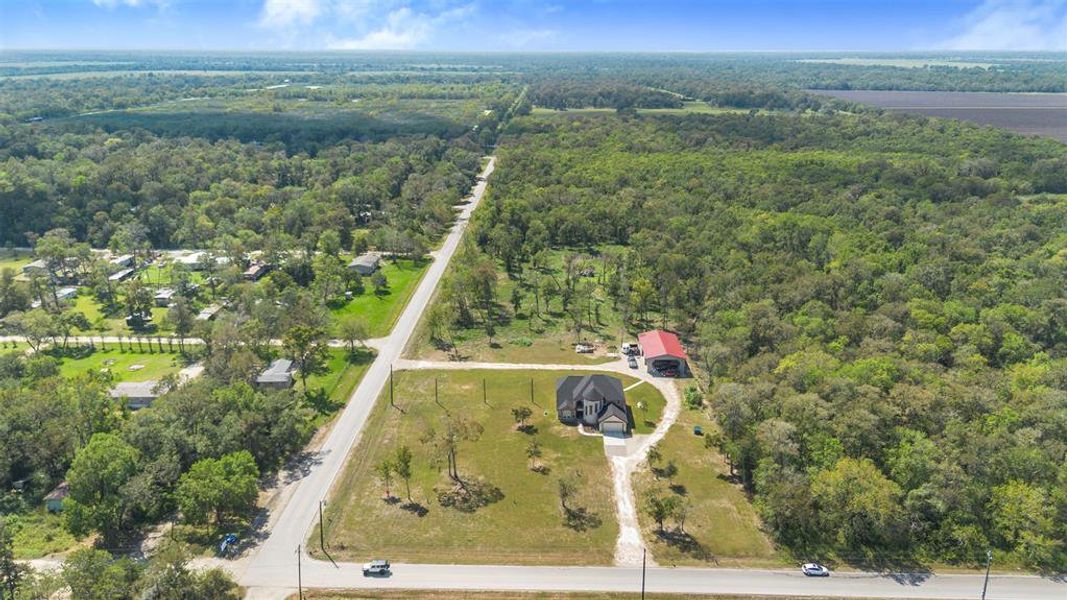 This aerial photo showcases a spacious rural property surrounded by lush greenery and trees, featuring a modern house with a large driveway and a sizable outbuilding. The property offers privacy and tranquility, with nearby roads providing easy access.