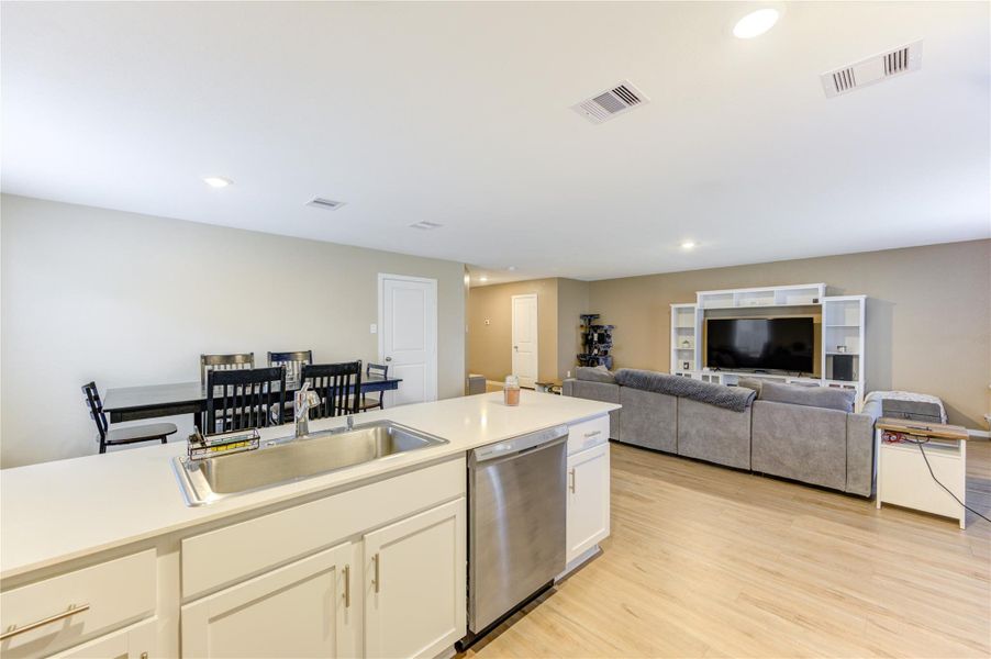 This open-concept living space features a modern kitchen with a central island and stainless steel appliances, seamlessly flowing into a cozy living area with a large sectional sofa and entertainment unit. The dining area is adjacent, creating a perfect setup for entertaining. Light vinyl plank flooring and recessed lighting enhance the bright, spacious feel.