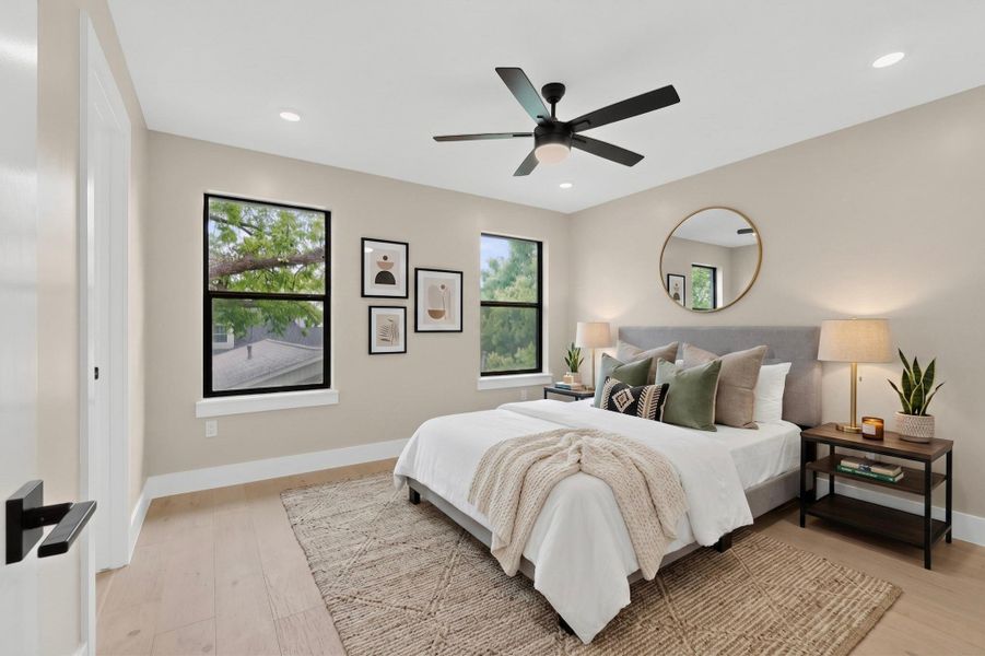 A tranquil primary bedroom features light wood flooring, a ceiling fan, and natural light from several windows.