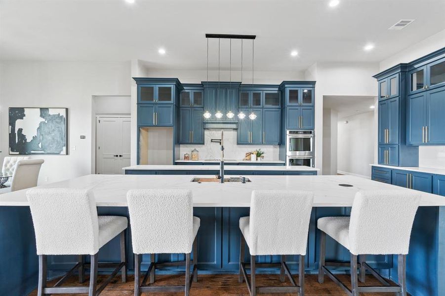 Kitchen featuring stainless steel double oven, blue cabinetry, backsplash, decorative light fixtures, and a kitchen island with sink