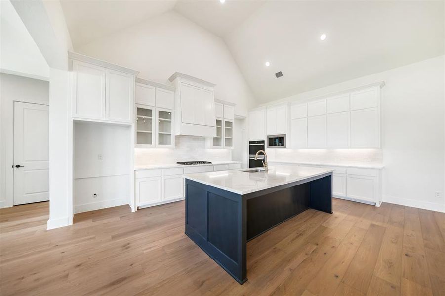 Kitchen featuring white cabinetry, backsplash, glass insert cabinets, an island with sink, and high vaulted ceiling