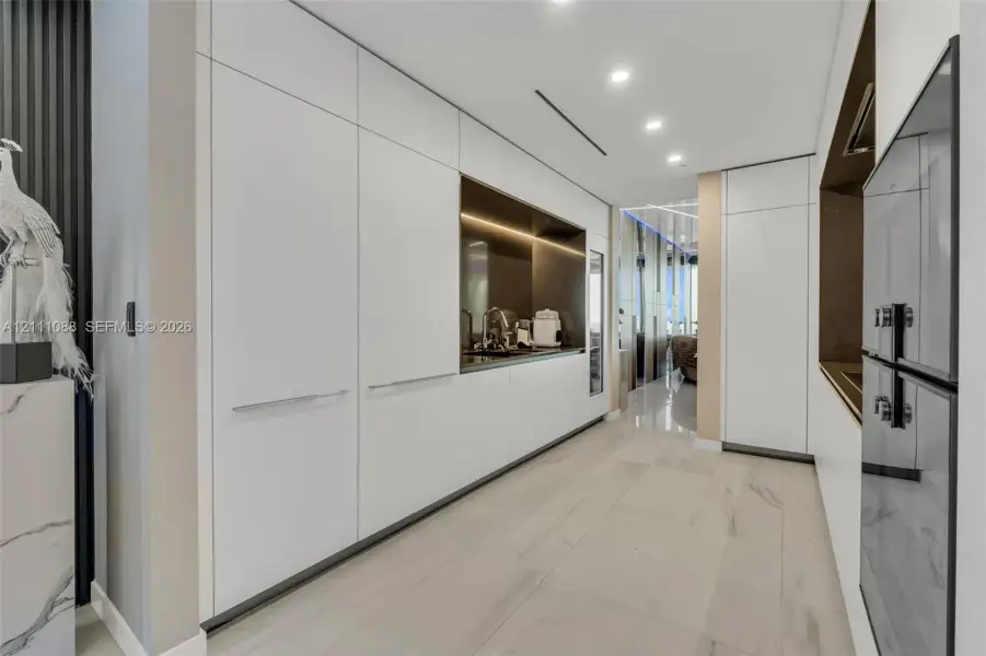 Sleek floor-to-ceiling white cabinetry, integrated recessed lighting, stone-finish tile flooring, built-in dual wall ovens, and a recessed metallic-finish backsplash Sleek floor-to-ceiling white cabinetry, integrated recessed lighting, stone-finish tile flooring, built-in dual wall ovens, and a recessed metallic-finish backsplash