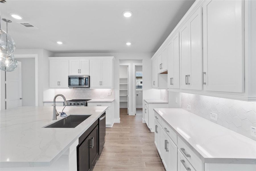 The open-concept gourmet kitchen designed for both cooking and entertaining. It features elegant white cabinetry, pristine quartz countertops, and a large central island that doubles as a functional breakfast bar. The cooking area is a chef's dream with a sleek stainless steel gas range with oven, and microwave hood vent, all complemented by a chic white tile backsplash.