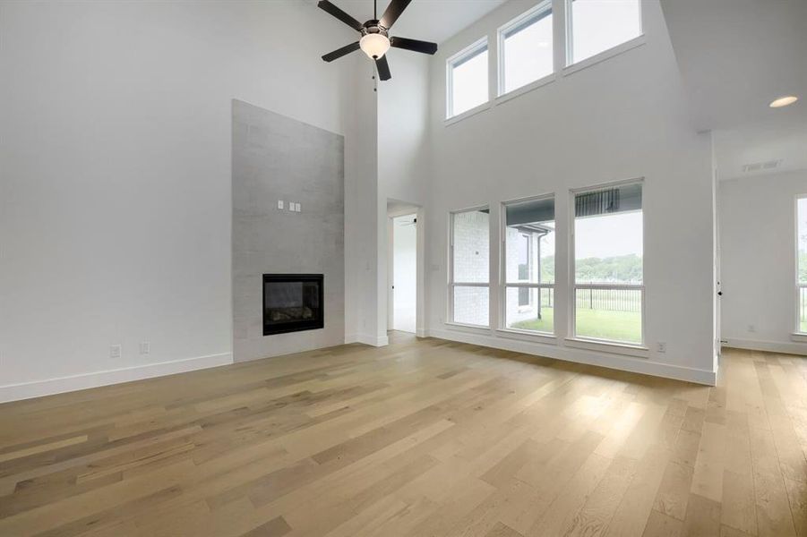 Two-story living area featuring a modern fireplace with a light-toned surround, wood-finish flooring, a ceiling fan, and multiple large windows Two-story living area featuring a modern fireplace with a light-toned surround, wood-finish flooring, a ceiling fan, and multiple large windows