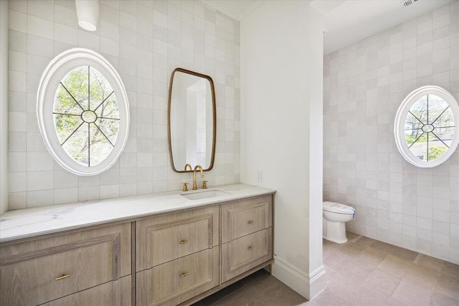 This secondary bathroom features tiled walls and flooring, beautiful oval windows, and a porcelain under-mount sink with gold fixtures.