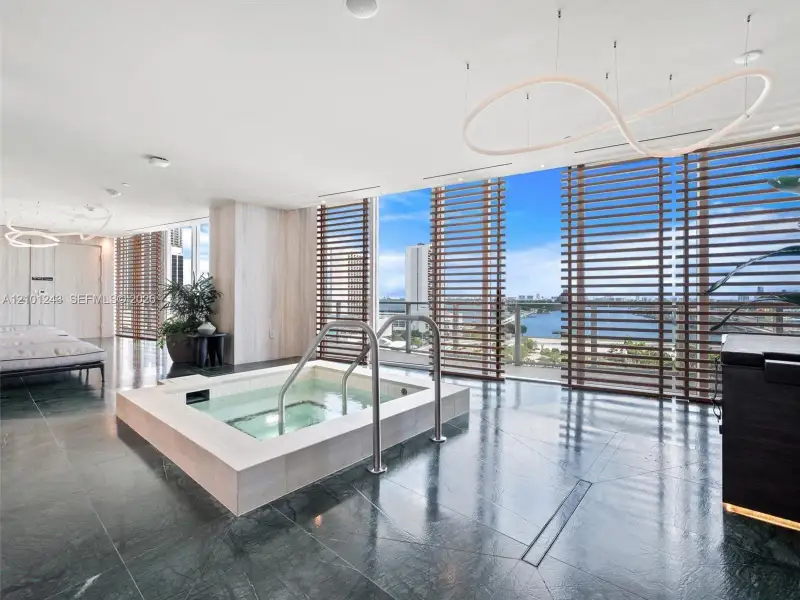 Built-in soaking tub with light-toned stone surround, polished dark stone flooring, floor-to-ceiling windows with horizontal wood-finish louvers, and architectural pendant lighting Built-in soaking tub with light-toned stone surround, polished dark stone flooring, floor-to-ceiling windows with horizontal wood-finish louvers, and architectural pendant lighting