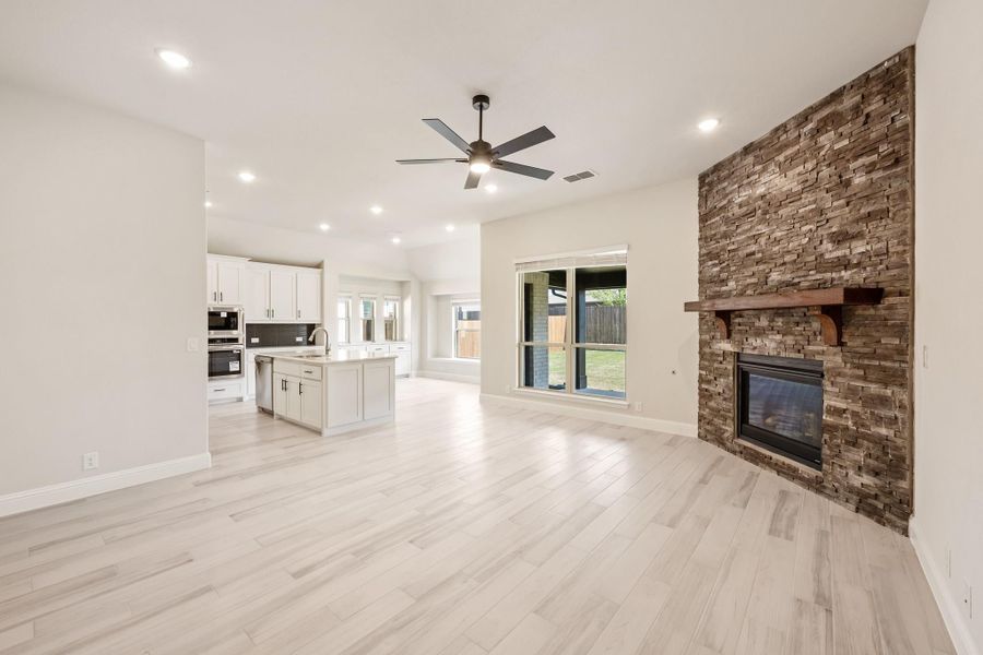 Open living area with stacked stone fireplace, ceiling fan, and kitchen with white cabinets and island in background Open living area with stacked stone fireplace, ceiling fan, and kitchen with white cabinets and island in background