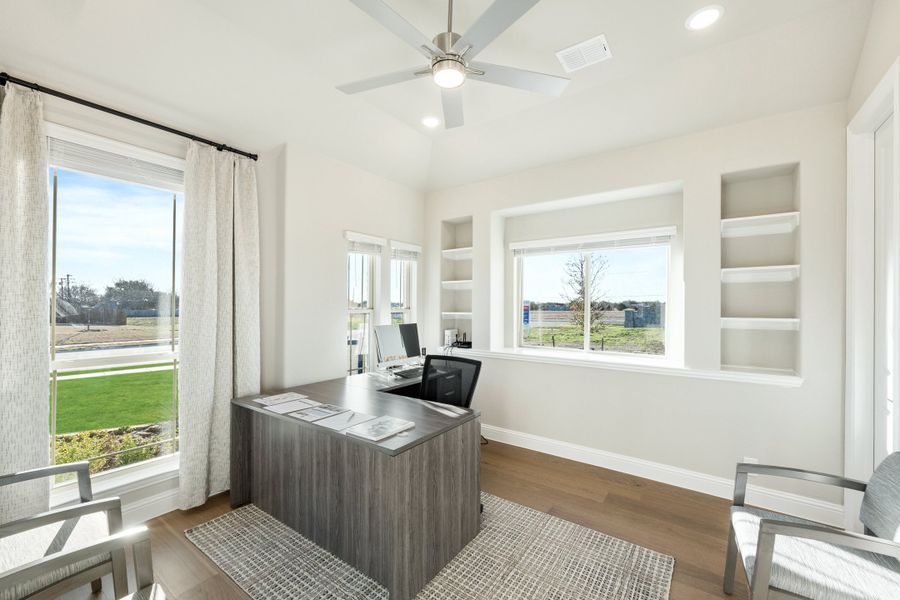Home office with dark wood desk, built-in shelves, ceiling fan, and large windows with natural light Home office with dark wood desk, built-in shelves, ceiling fan, and large windows with natural light
