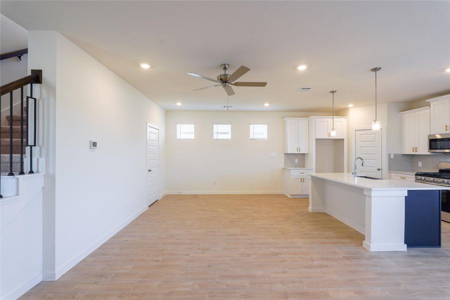 This modern, open-concept space features a bright kitchen with white cabinetry, stainless steel appliances, and a large island with pendant lighting. The living area has wood flooring, recessed lighting, and a ceiling fan. A staircase with a stylish railing leads to the upper level, adding a touch of elegance.