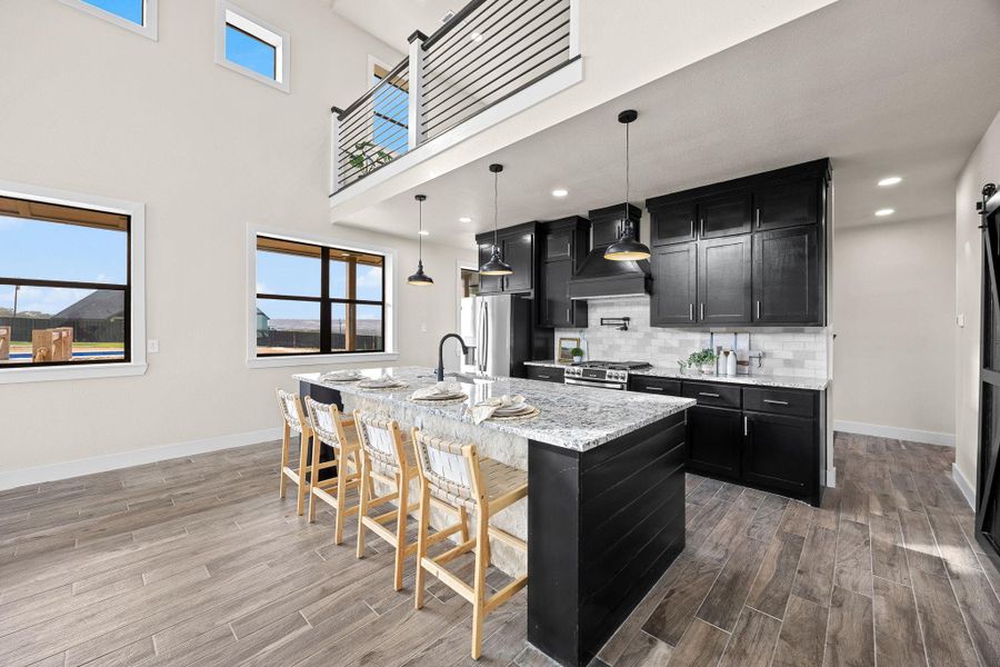 Kitchen featuring dark cabinetry, a barn door, tasteful backsplash, a center island with sink, and light stone countertops Kitchen featuring dark cabinetry, a barn door, tasteful backsplash, a center island with sink, and light stone countertops