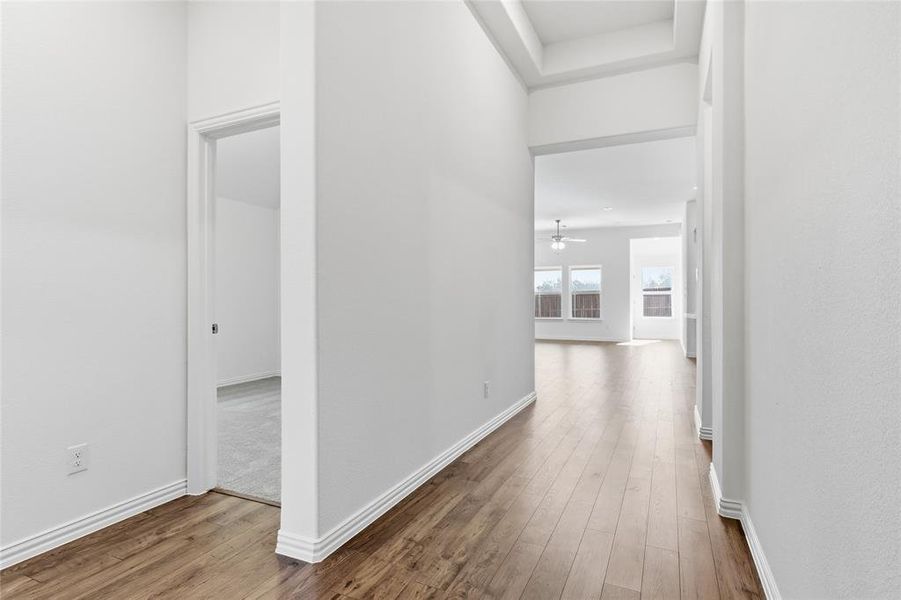 Hallway featuring wood-finish flooring, white baseboards, a tray ceiling, and a framed doorway leading to a carpeted room