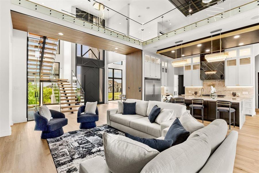 Open-concept interior featuring light wood-finish flooring, a contemporary staircase with glass railings, a chef's kitchen with a large island and pendant lighting, a two-story foyer with a dark paneled door, and a second-story interior balcony