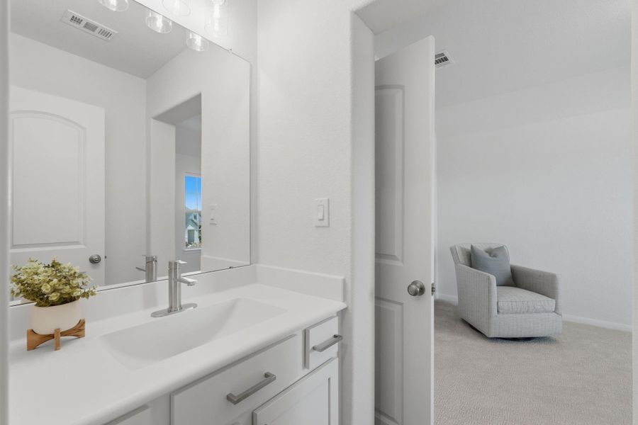 This photo showcases a bright, modern bathroom with a sleek white vanity and a large mirror. Adjacent is a glimpse into a cozy, carpeted room featuring a comfortable armchair.