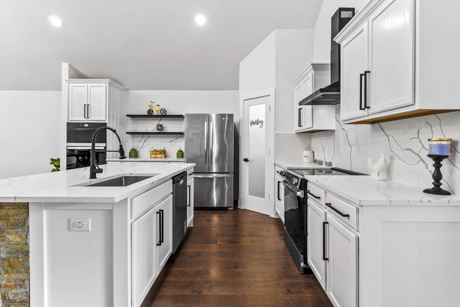 This modern kitchen features sleek white cabinetry, quartz countertop, and a spacious island with a black faucet. It includes stainless steel appliances, a pantry with a frosted glass door, and stylish dark wood flooring.