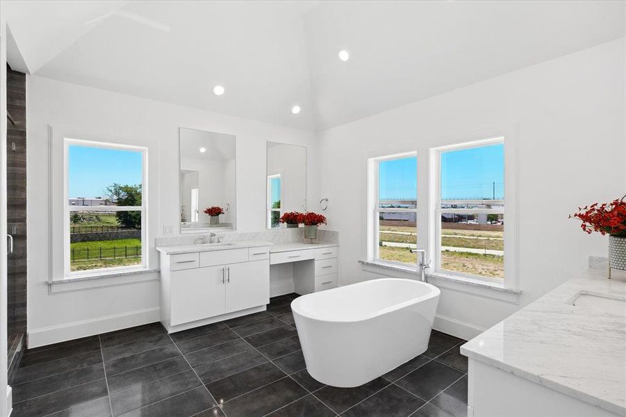 Spacious bathroom featuring a freestanding tub, dual vanities with white cabinetry and light-toned countertops, dark tile flooring, and a vaulted ceiling with recessed lighting
