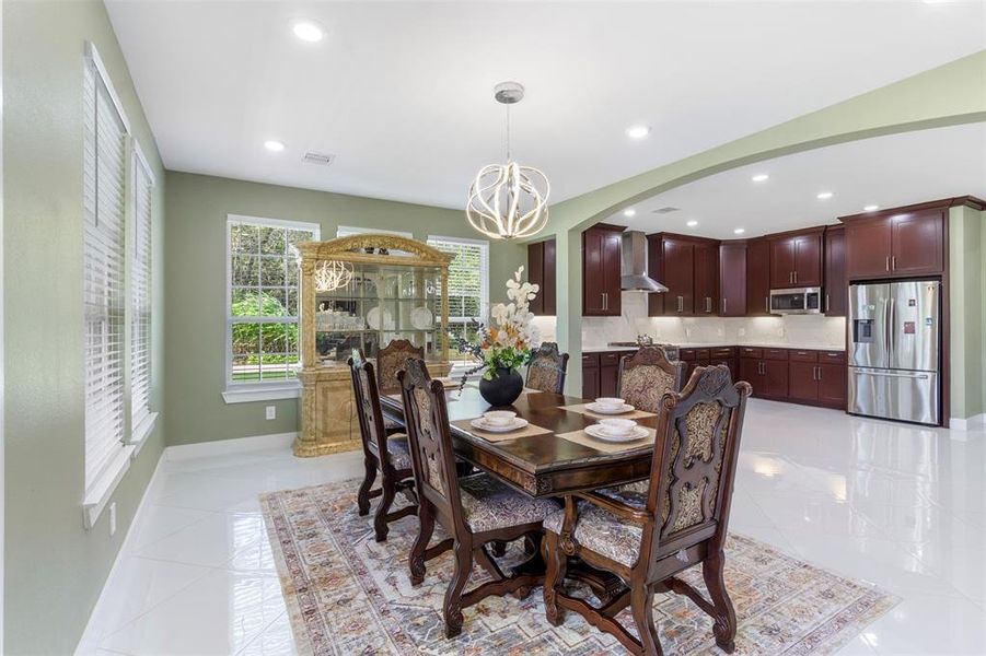 This photo showcases a spacious, open-concept dining and kitchen area. The dining space features elegant, ornate chairs and a large wooden table, complemented by a stylish light fixture. The kitchen boasts dark wood cabinetry, modern stainless steel appliances, and ample counter space. Large windows offer natural light, enhancing the room's bright and inviting atmosphere.