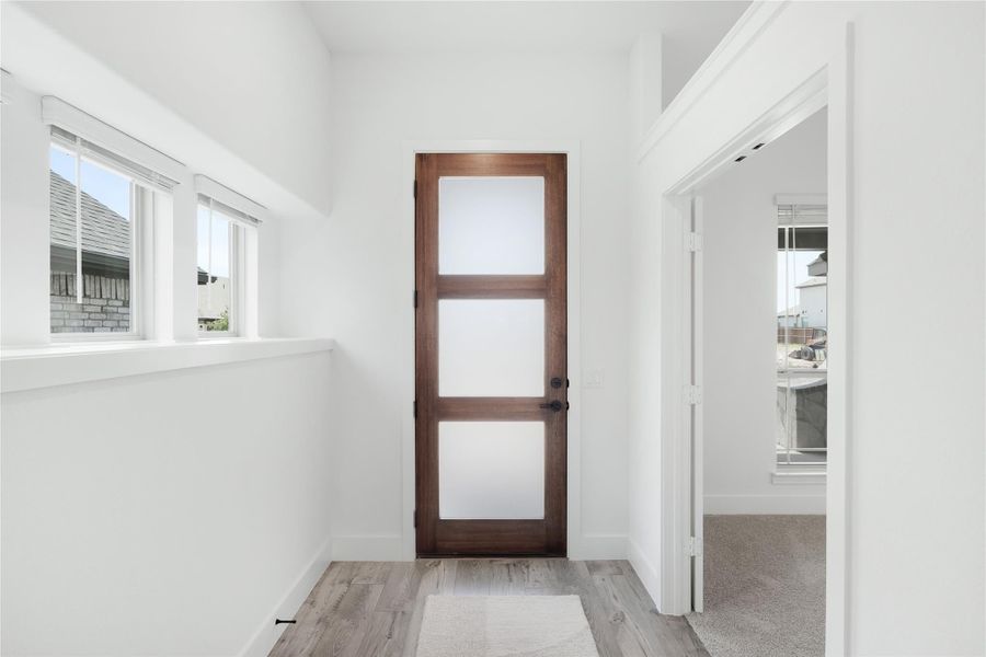 Hallway with dark wood door featuring frosted glass panels, white walls, and light hardwood floors Hallway with dark wood door featuring frosted glass panels, white walls, and light hardwood floors