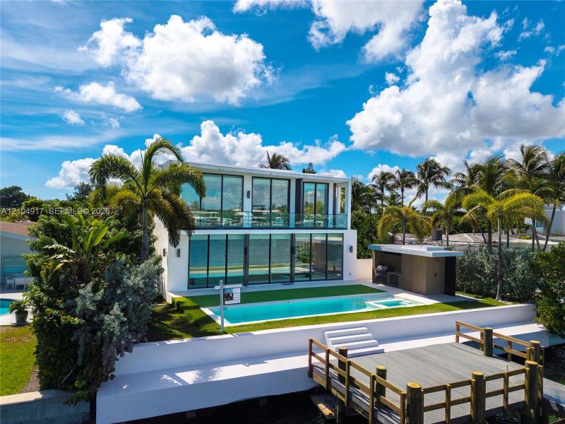 Two-story modern residence featuring floor-to-ceiling glass wall panels, an in-ground swimming pool, a built-in outdoor kitchen structure, and a wood-finish dock