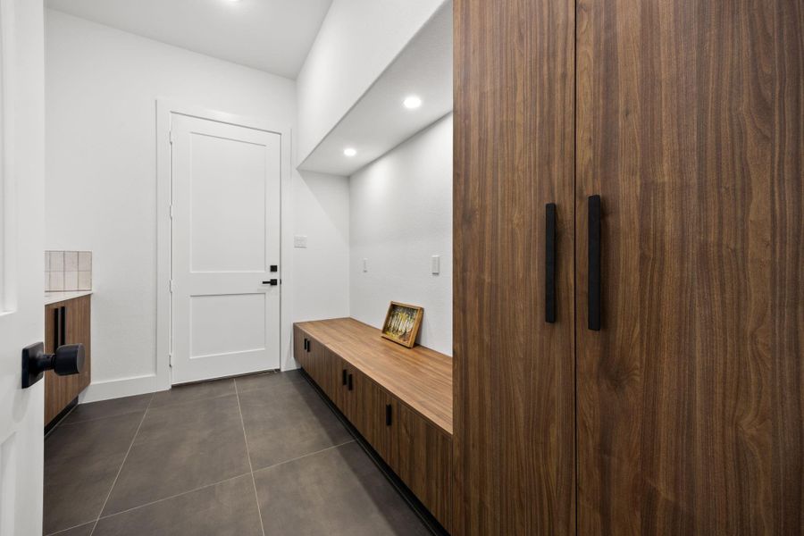 This photo showcases a modern mudroom with sleek, dark tile flooring, white walls, and stylish wood cabinetry. It features built-in storage with a bench, recessed lighting, and a clean, minimalist design.