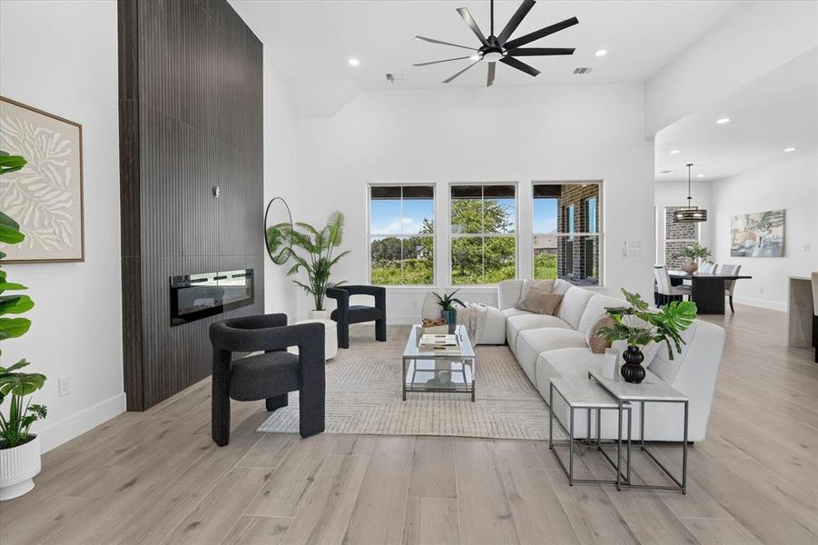 Spacious living area featuring a tall vaulted ceiling, wood-finish flooring, a contemporary fireplace with a dark paneled surround, an oversized ceiling fan, and large windows providing abundant natural light