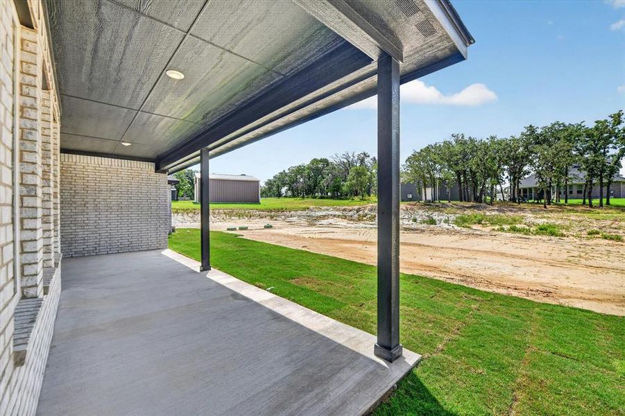 Covered concrete patio with recessed lighting, supported by dark posts, featuring a light brick exterior and an expanse of green lawn Covered concrete patio with recessed lighting, supported by dark posts, featuring a light brick exterior and an expanse of green lawn
