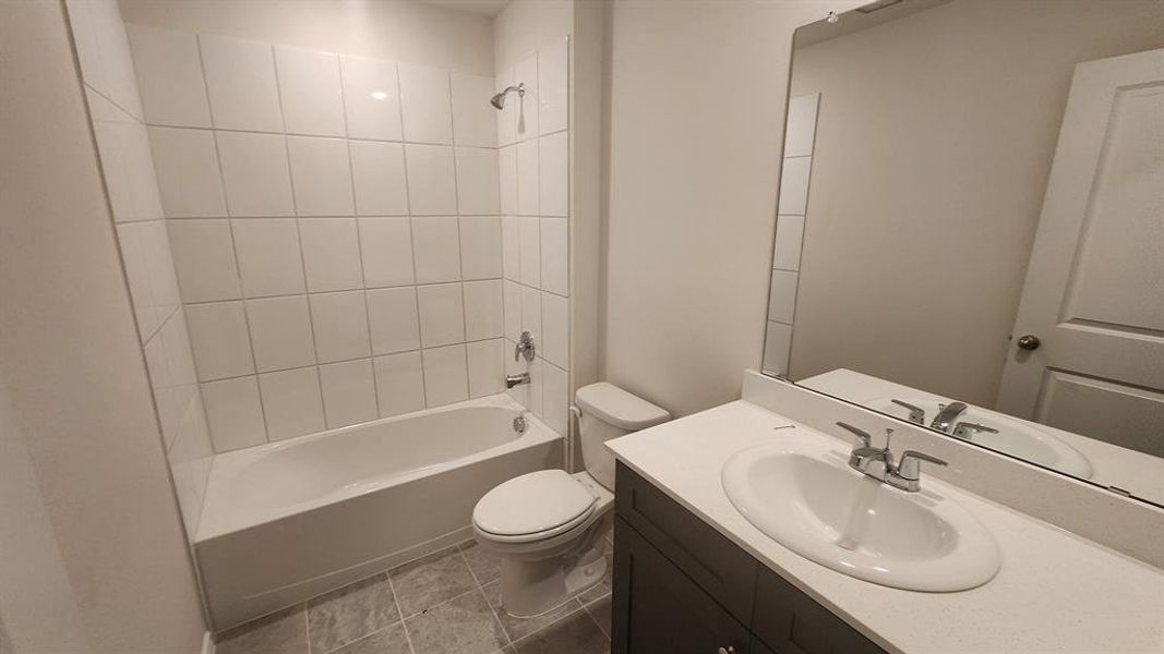 Full bathroom featuring a white tub with tile surround, a built-in toilet, a vanity with a white countertop and integrated sink, a large wall mirror, and grey floor tiles Full bathroom featuring a white tub with tile surround, a built-in toilet, a vanity with a white countertop and integrated sink, a large wall mirror, and grey floor tiles