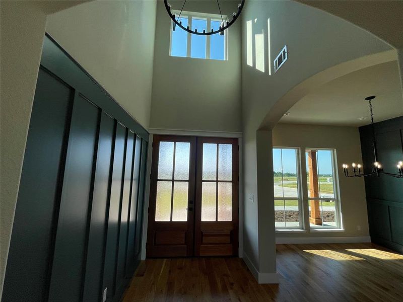 Grand entry featuring double doors with frosted glass panels, wood-finish flooring, a two-story ceiling, an overhead chandelier, and a decorative wall paneling accent