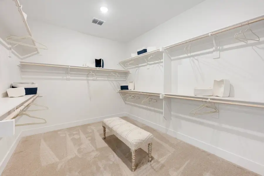 Walk-in closet with white shelving, hanging rods, carpet flooring, and a small upholstered bench in the center. Walk-in closet with white shelving, hanging rods, carpet flooring, and a small upholstered bench in the center.