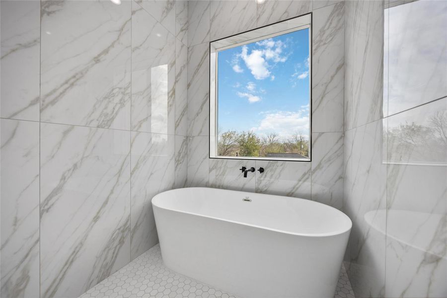This luxurious spa-like retreat features a sleek freestanding soaking tub set against floor-to-ceiling marble-style tile, beautifully framed by a large picture window that fills the space with natural light and serene outdoor views.