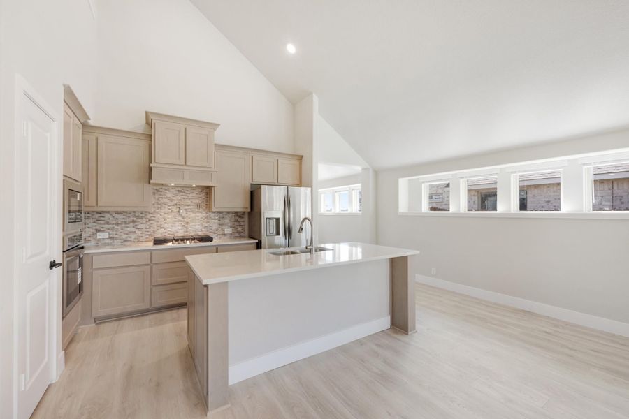Open kitchen with large center island, beige cabinets, tile backsplash, and light hardwood floors under vaulted ceiling