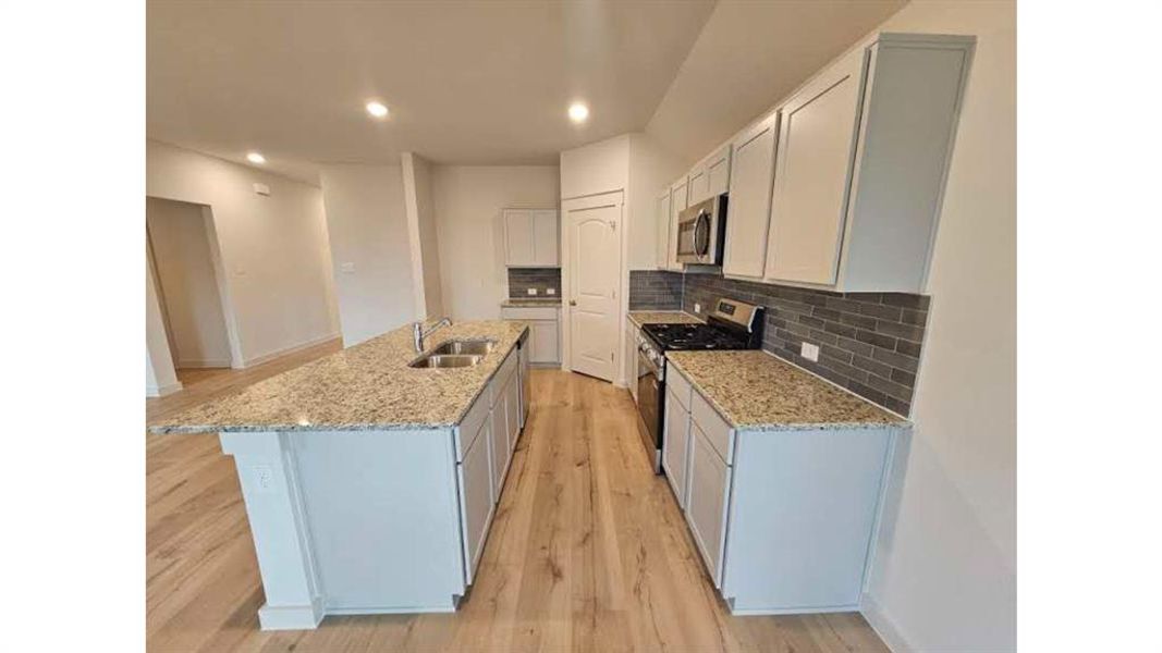 Kitchen featuring a large central island with double sinks, light-toned granite countertops, light wood-finish flooring, white cabinetry, and stainless steel appliances
