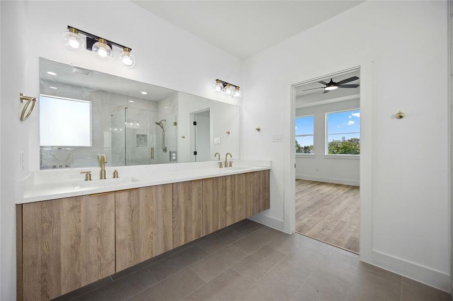 Modern bathroom with a double vanity, gold fixtures, oversized mirror, and a walk-in shower. Enjoy the clean, bright design with tile flooring and private access from the primary suite.