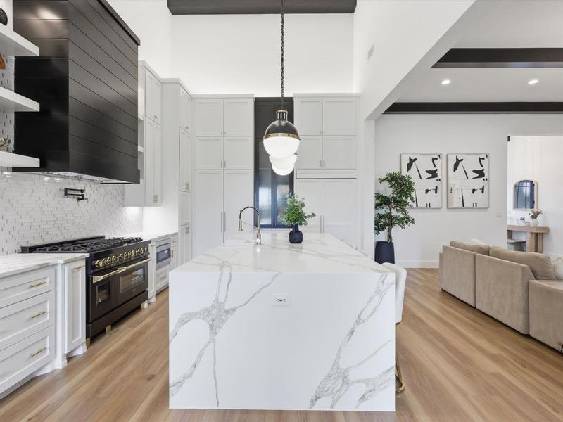 Open-concept kitchen featuring a waterfall island with integrated sink, white cabinetry, and a black range with custom hood