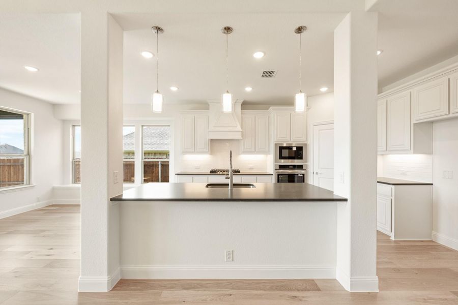 Open kitchen with white cabinets, dark countertop island, pendant lights, and stainless steel appliances on light hardwood floors Open kitchen with white cabinets, dark countertop island, pendant lights, and stainless steel appliances on light hardwood floors