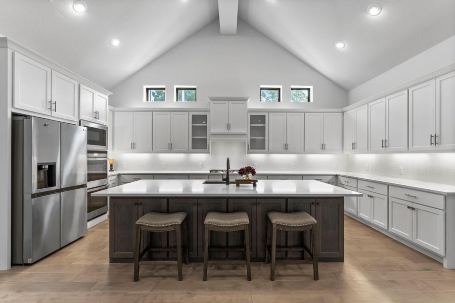 This is a head-on shot of the kitchen. It features white upper and lower cabinets with dark hardware, a white tile backsplash, and a large center island with a dark wood base and a white countertop. Three bar stools (not included in the sale) are tucked under the island. A stainless steel refrigerator and double oven are on the left. This is a head-on shot of the kitchen. It features white upper and lower cabinets with dark hardware, a white tile backsplash, and a large center island with a dark wood base and a white countertop. Three bar stools (not included in the sale) are tucked under the island. A stainless steel refrigerator and double oven are on the left.