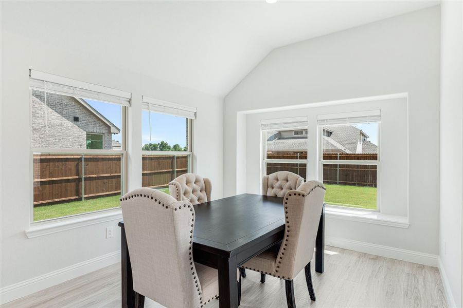 Dining area with dark wood table, four upholstered chairs, vaulted ceiling, and large windows overlooking backyard