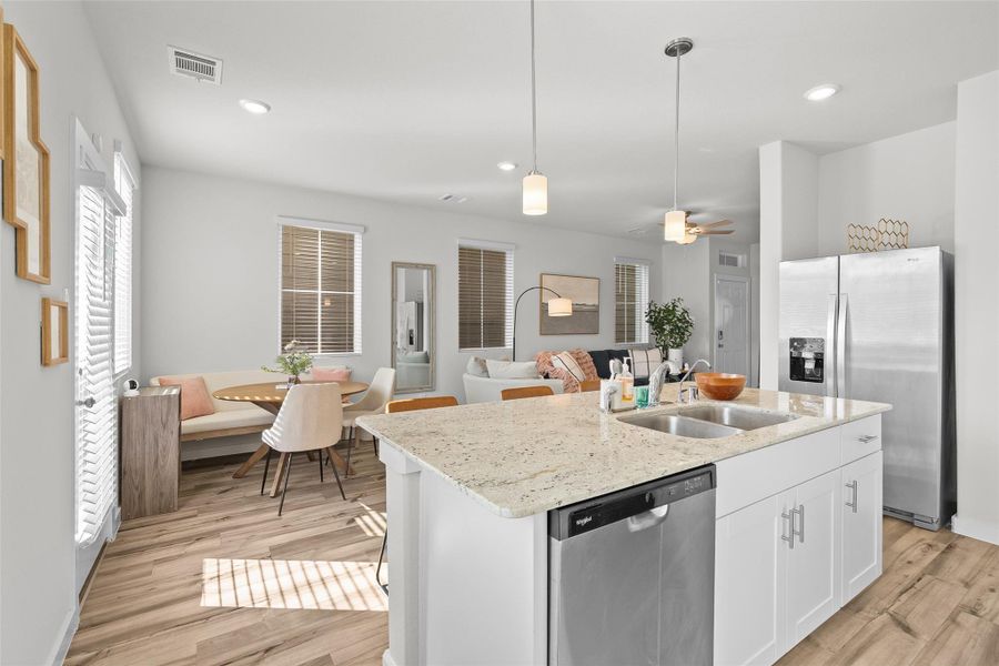 This photo showcases a bright, open-concept living space featuring a modern kitchen with granite countertops, stainless steel appliances, and a central island with a sink. Adjacent to the kitchen is a cozy dining area with a stylish table and chairs. The space is warmly lit with natural light, accentuated by contemporary light fixtures, and has a comfortable seating area perfect for relaxation.