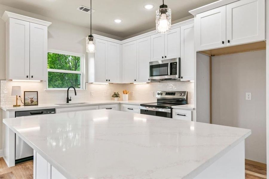 Kitchen featuring a large central island with a light-toned countertop, white cabinetry with black hardware, stainless steel appliances, and pendant lighting