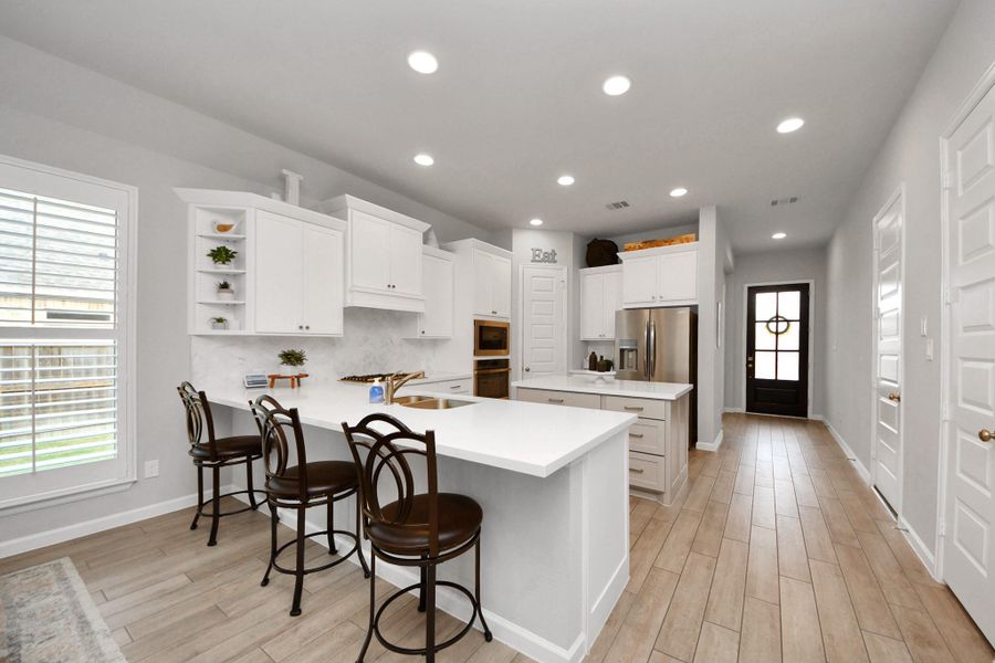 This modern kitchen features white cabinetry, a spacious island with seating for three, and stainless steel appliances. It has wood-style flooring, recessed lighting, and a large window for natural light. The open layout leads to a back door, enhancing accessibility and flow. This modern kitchen features white cabinetry, a spacious island with seating for three, and stainless steel appliances. It has wood-style flooring, recessed lighting, and a large window for natural light. The open layout leads to a back door, enhancing accessibility and flow.