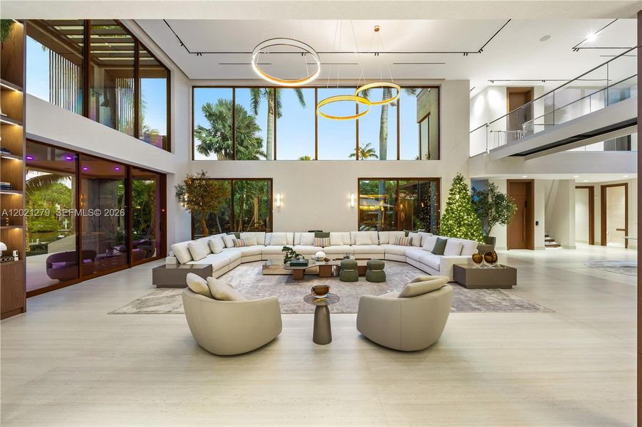 Grand double-height living room with wraparound sectional seating, light stone floors, dual ring chandelier, full-wall glass overlooking tropical waterfront, open second-floor walkway with glass railing, and adjacent open-plan areas.