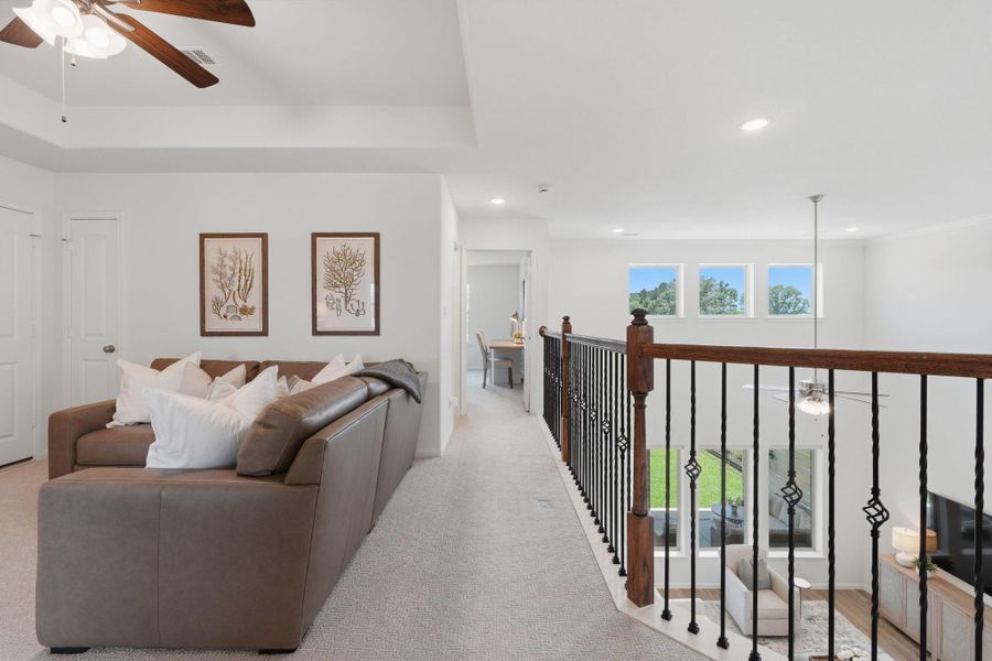 This photo showcases a bright, open upstairs landing area with a cozy seating nook, decorative wall art, and elegant railing overlooking the living space below. Large windows provide plenty of natural light, and there's a ceiling fan for added comfort.