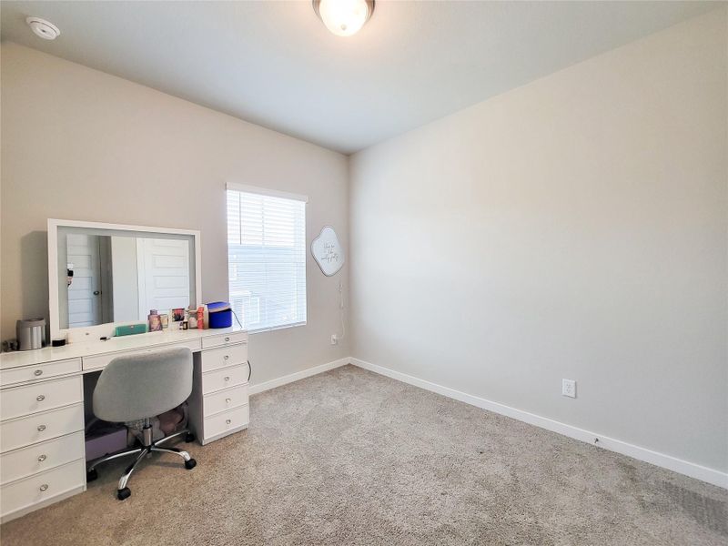 Bright secondary bedroom, carpeted room with neutral-color walls, featuring a window for natural light. There's a white vanity with a mirror and chair, offering a versatile space suitable for a bedroom or office.
