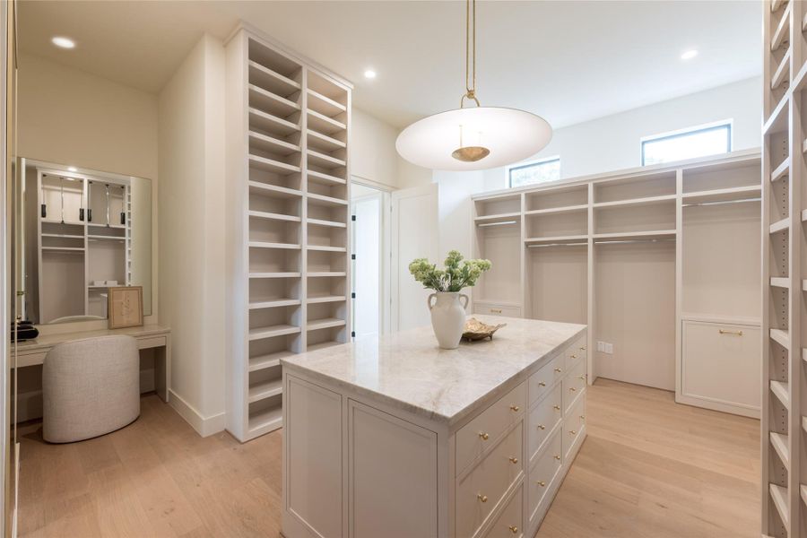 This spacious primary closet features custom built-ins, a marble-topped island with brass hardware, and dedicated vanity space for added functionality. Natural light pours in through clerestory windows, highlighting the soft oak floors and refined millwork. This spacious primary closet features custom built-ins, a marble-topped island with brass hardware, and dedicated vanity space for added functionality. Natural light pours in through clerestory windows, highlighting the soft oak floors and refined millwork.