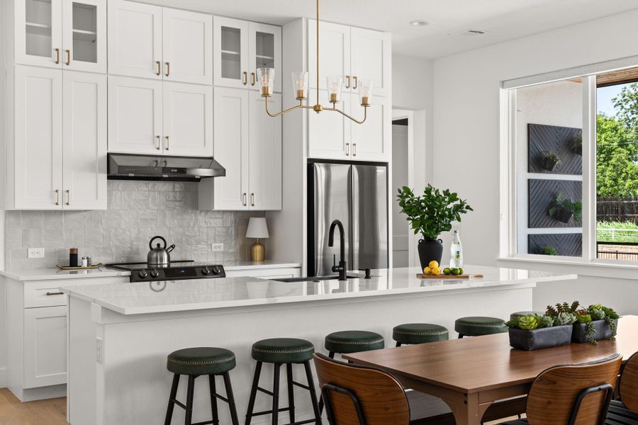 This modern kitchen features sleek white cabinetry, a stainless steel fridge, and a stylish black range hood. The spacious island offers plenty of seating complemented by a chic chandelier light above. Large windows provide ample natural light, enhancing the bright and airy feel.