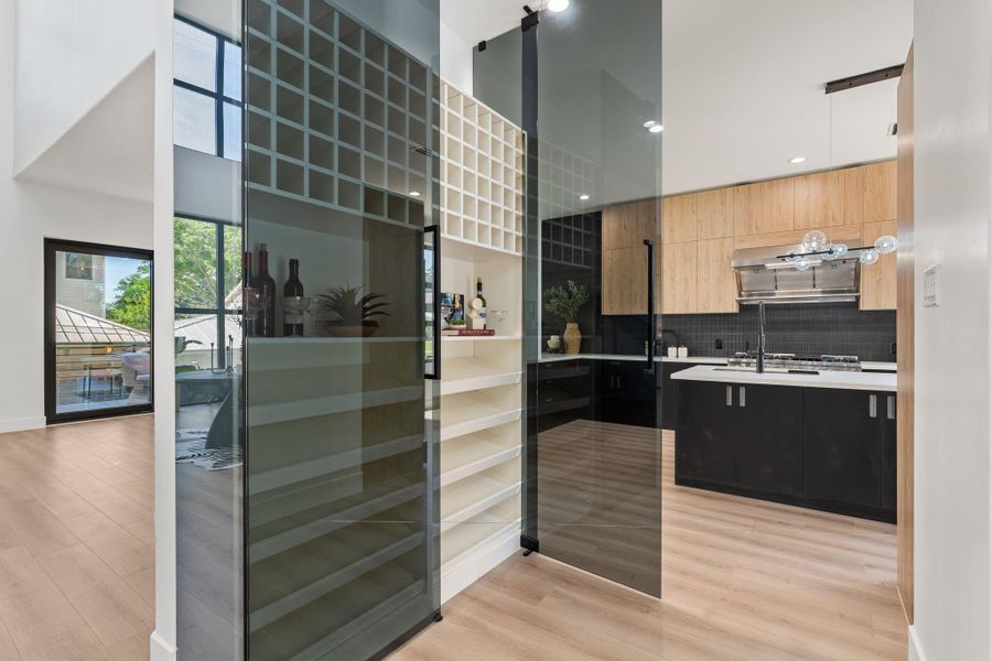 The smoked glass wine wall is one of the home's most distinctive architectural moments — a floor-to-ceiling grid of frosted glass panels with wine storage visible within, positioned as a transparent divider between the entry and the kitchen. Functional and striking in equal measure.