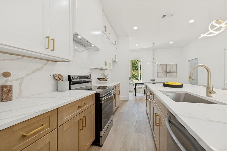Modern kitchen featuring an expansive island with an undermount sink and a gold-tone faucet, white upper cabinetry, and wood-finish lower cabinetry Modern kitchen featuring an expansive island with an undermount sink and a gold-tone faucet, white upper cabinetry, and wood-finish lower cabinetry