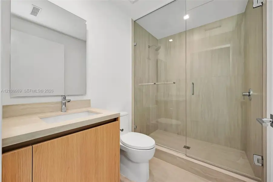Wood-finish cabinetry with integrated countertop and rectangular sink, polished chrome fixtures, frameless glass shower enclosure, and full-height stone-finish tile surround