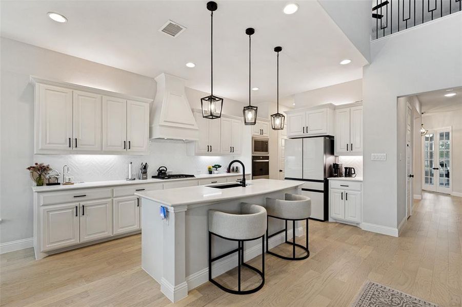 Spacious kitchen featuring an expansive island with seating, a professional range hood, white shaker cabinetry with dark hardware, a white herringbone tile backsplash, and light wood-finish flooring