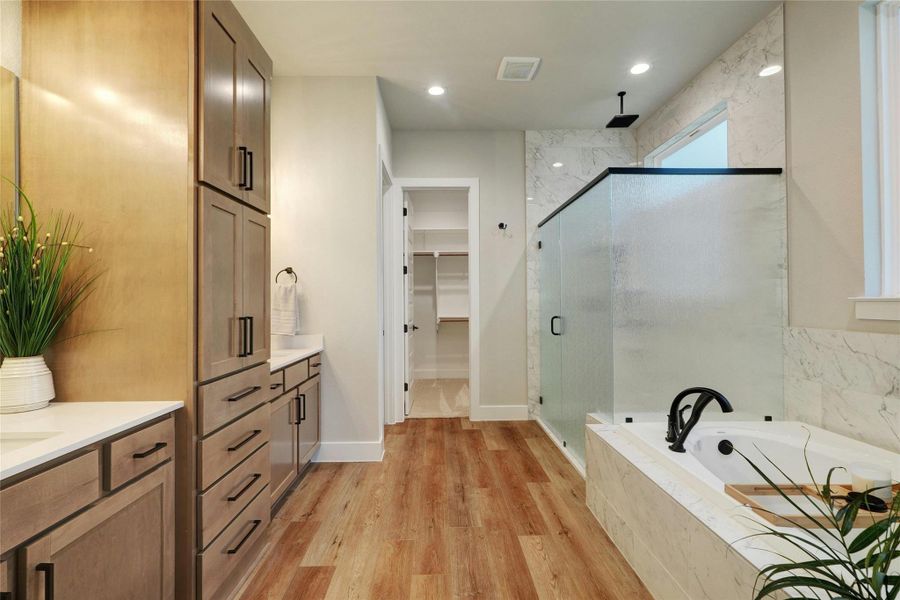 Contemporary bathroom featuring a spacious walk-in shower with frosted glass and a rainfall showerhead, a large soaking tub with matte black fixtures, extensive cabinetry with dark hardware, dual vanity sinks with light countertops, and wood-finish flooring